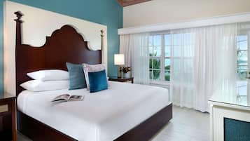 Una cama grande con cabecera de madera, ropa de cama blanca y almohada azul. Hay una mesita de noche con una lámpara y una pequeña mesa junto a la cama. Se ve una ventana con cortinas blancas y una planta sobre la mesita de noche.