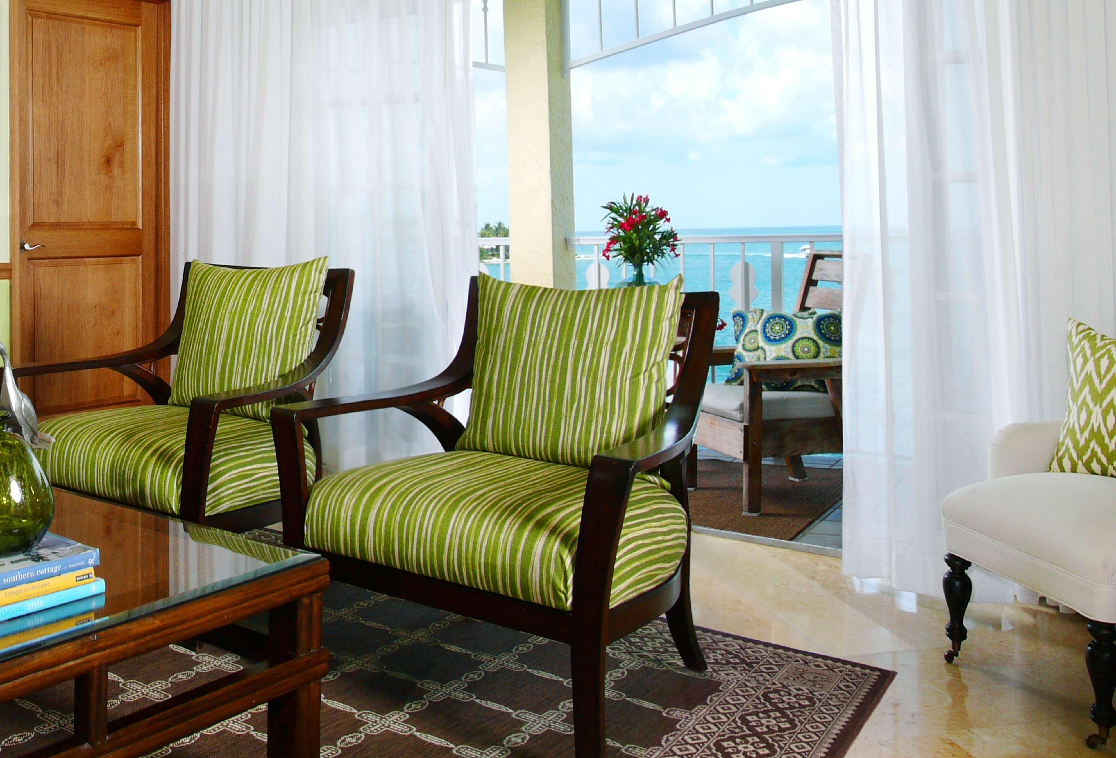 A living room with two striped armchairs, a glass coffee table, and a view of the ocean through a balcony.