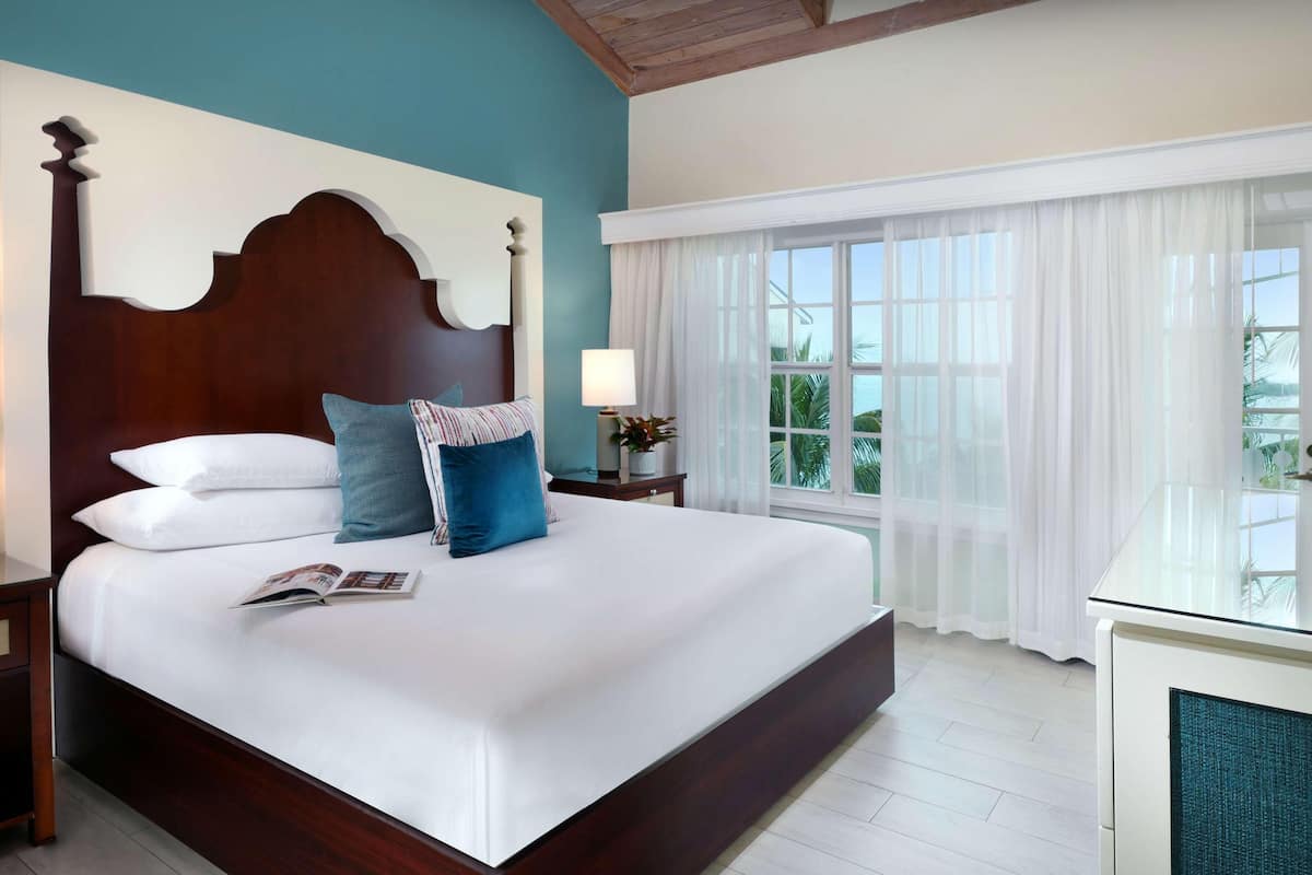 A large bed with a wooden headboard, white bedding, and a blue pillow. There is a nightstand with a lamp and a small table beside the bed. A window with white curtains is visible, and a plant is on the nightstand.