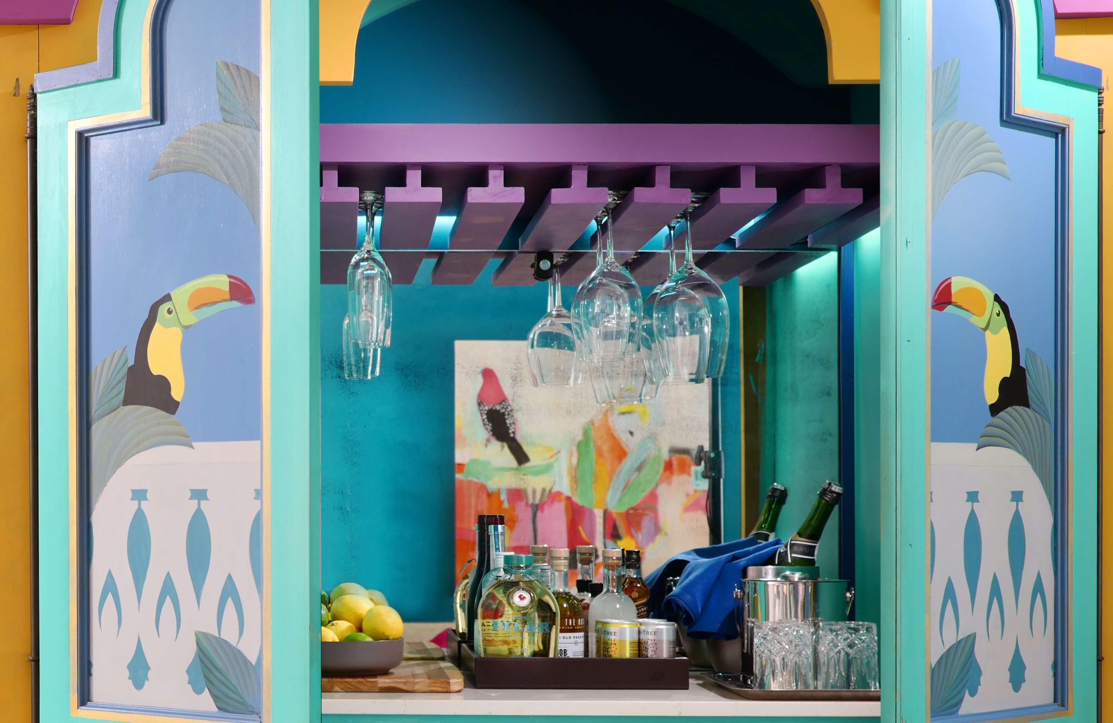 Zona de bar con murales coloridos, un frutero y diferentes botellas y vasos.