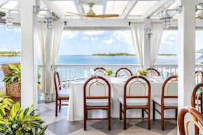An outdoor dining area with white tablecloths, wooden chairs, and a view of the ocean.