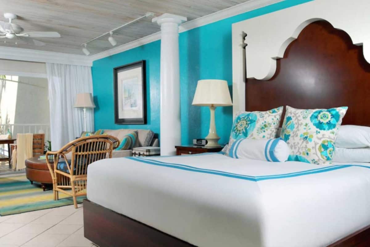 A hotel room with a large bed, a wooden headboard, a nightstand with a lamp, a chair, and a view of the outdoors.