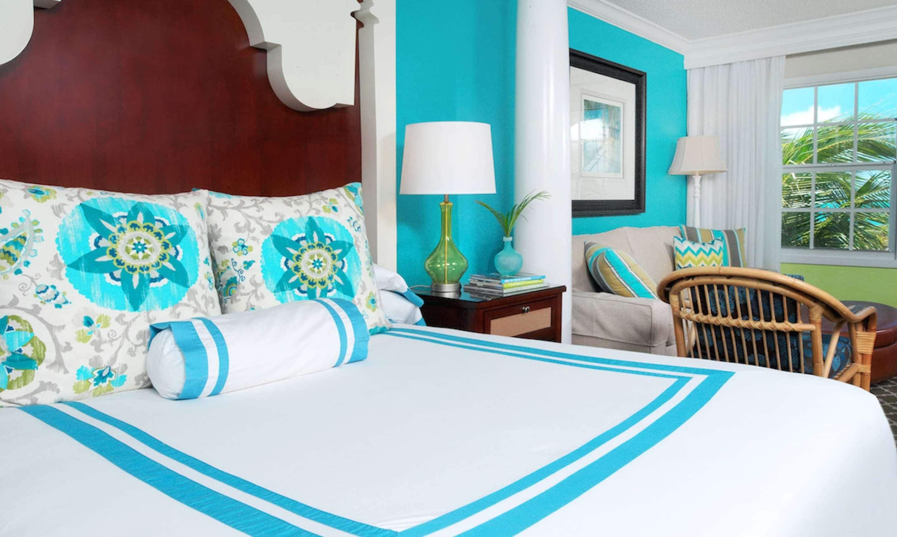 A hotel room with a large bed, a wooden headboard, a teal accent wall, a wicker chair, and a window with a view of palm trees.