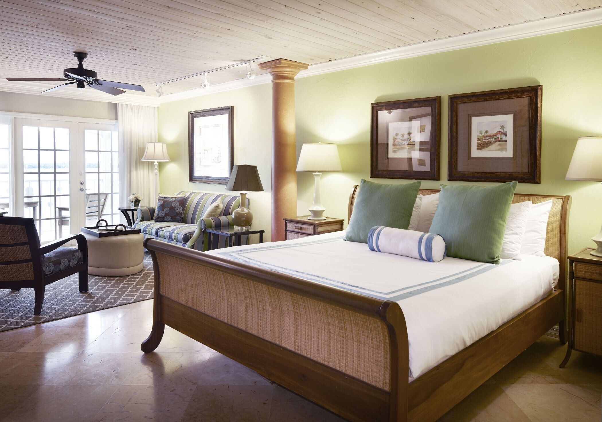 A spacious bedroom with a large bed, a sitting area with a sofa and armchairs, a ceiling fan, and framed pictures on the walls.