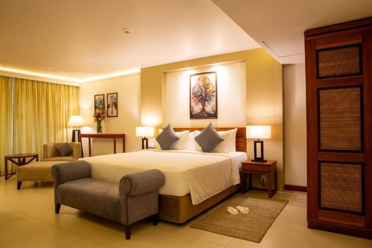 Habitación de hotel con una cama grande, dos mesitas de noche con lámparas, una silla, un escritorio y un amplio ventanal con cortinas.