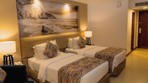 Habitación de hotel con dos camas, un cuadro grande de una playa, una mesita de noche con una lámpara y una puerta que da a otra habitación.