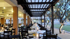 An outdoor dining area with tables set for two, covered by a pergola, and a view of a landscaped garden.