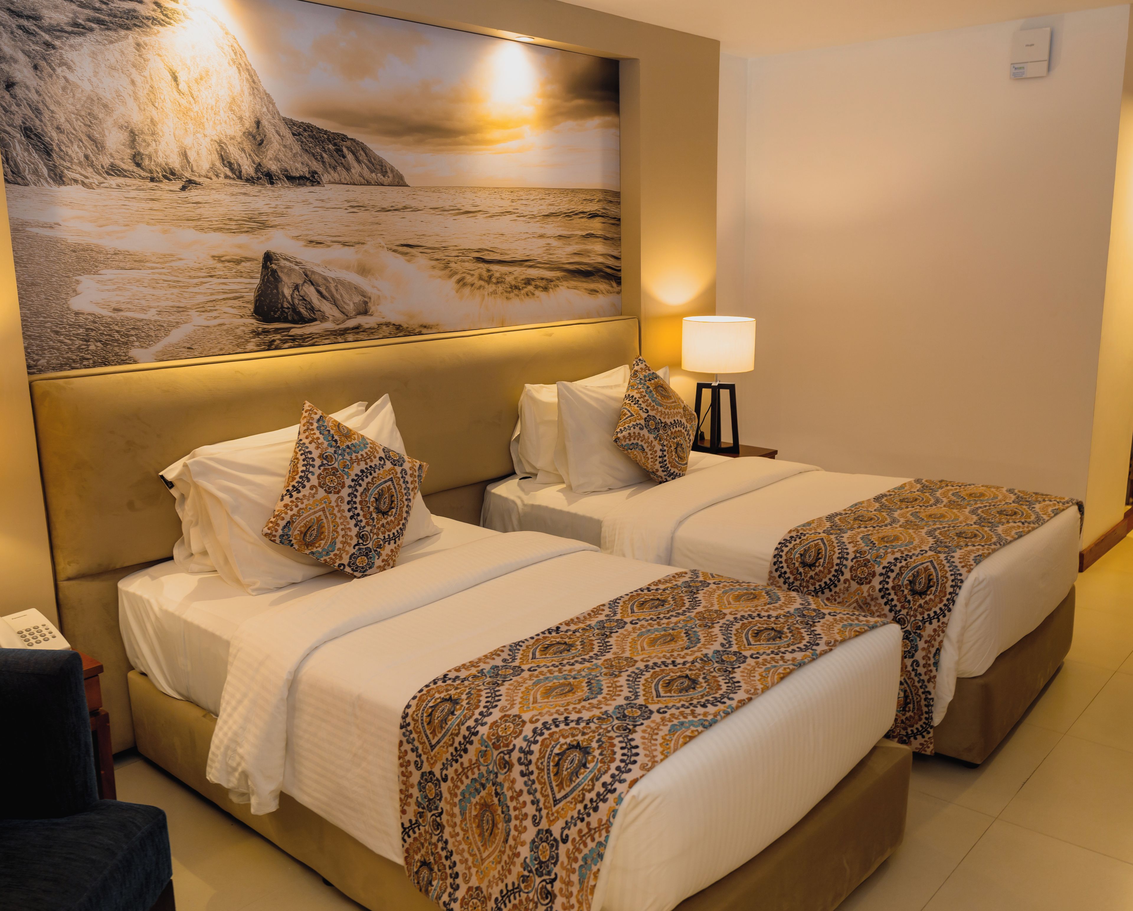 Una camera d'albergo con due letti, un quadro di grandi dimensioni raffigurante una costa, una lampada da comodino e un telefono sul comodino.
