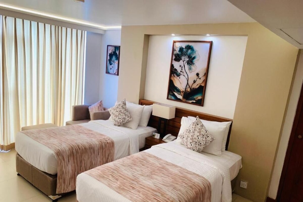 Habitación de hotel con dos camas, un ventanal con cortinas, un cuadro en la pared y una mesita de noche con lámpara.