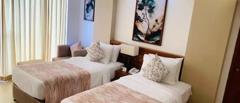 A hotel room with two beds, a large window with curtains, a painting on the wall, and a bedside table with a lamp.