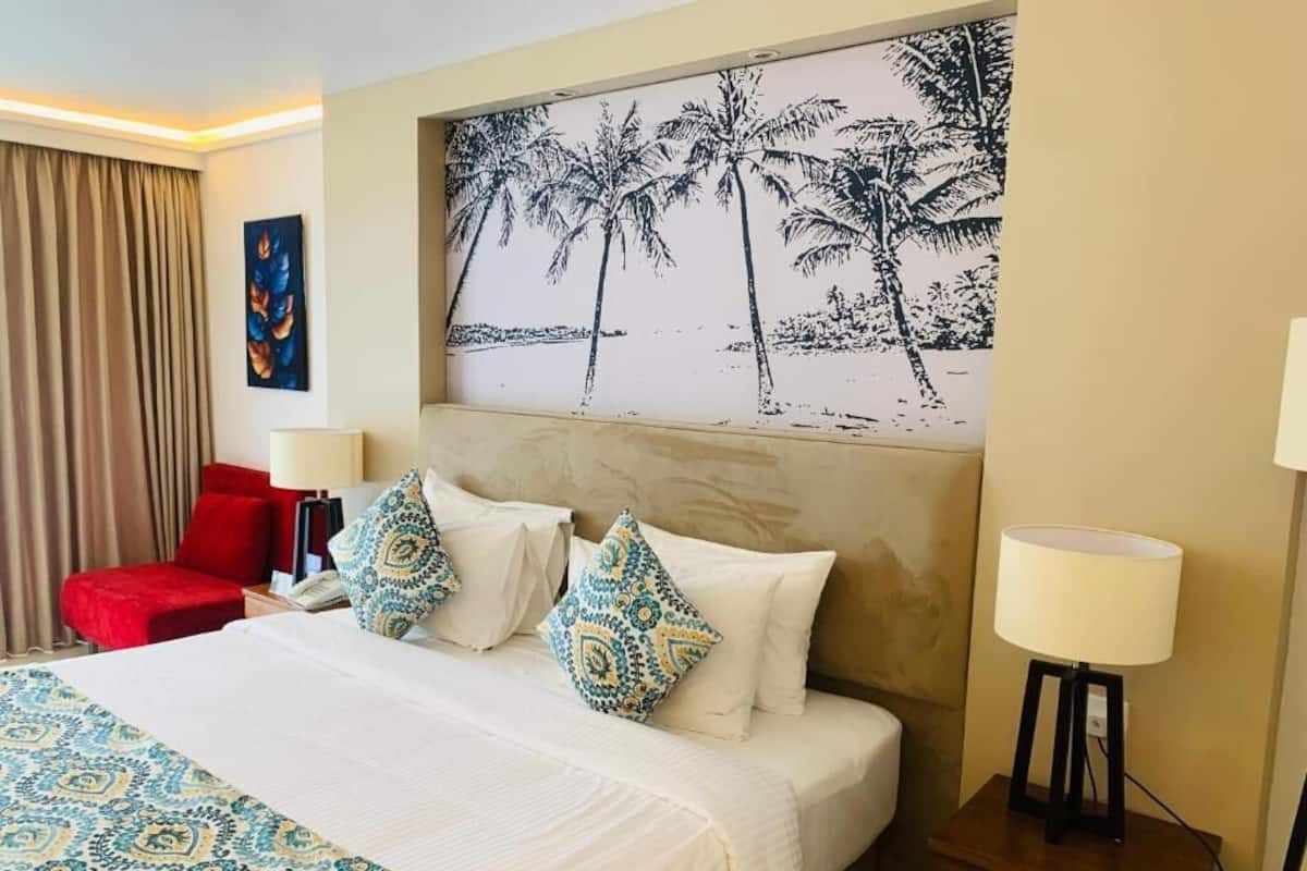 Una habitación de hotel con una cama, una silla roja, una mesita de noche, una lámpara y un cuadro de palmeras.