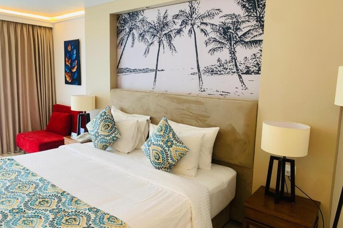 Quarto de hotel com cama, uma cadeira vermelha, mesa de cabeceira, abajur e pintura de palmeiras.