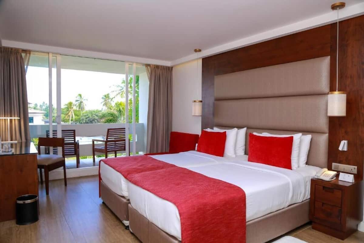 Habitación de hotel con una cama grande, un escritorio, sillas y un balcón con vistas a palmeras.