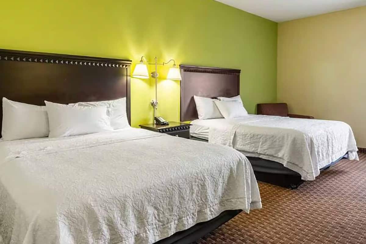 A hotel room with two beds, a nightstand, a lamp, and a chair.
