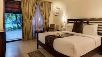 A hotel room with a bed, a desk, a chair, a window with curtains, and a view of greenery outside.