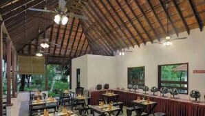 A restaurant with a high ceiling and wooden beams, tables set for dining, and large windows offering a view of greenery outside. - Abad Turtle Beach (Ambalapuzha)