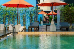 Un área de piscina con sillones de descanso y sombrillas, rodeada de plantas y un muro azul.