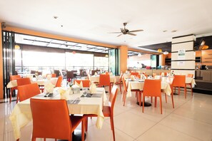 A spacious dining area with multiple tables and orange chairs, large windows, and a ceiling fan. - The Vogue Hotel, Pattaya (Pattaya)