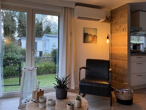 Ein modernes Wohnzimmer mit großem Fenster, einem schwarzen Ledersessel, einem runden Holztisch mit Kerzen und Pflanzen, und im Fenster erkennbarer Küchenbereich.