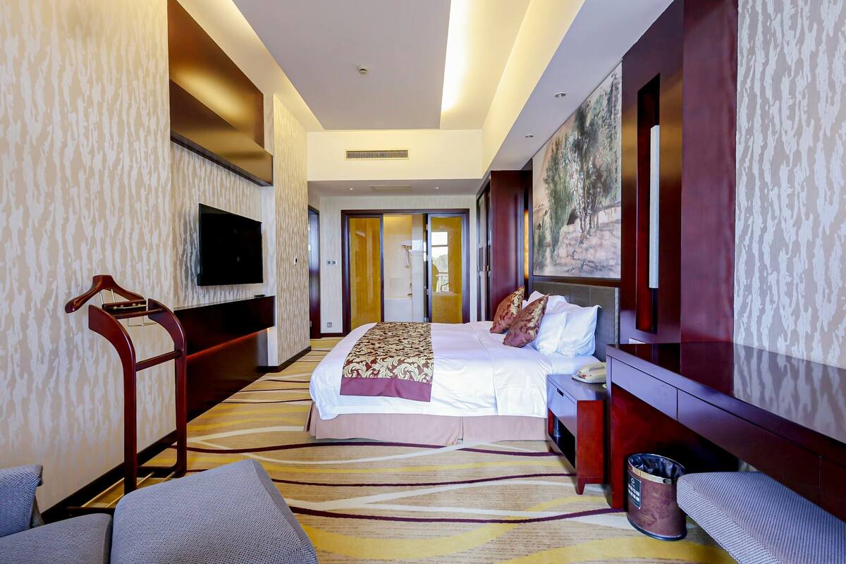 A hotel room with a large bed, a TV mounted on the wall, a wooden chair, a bench, and a painting on the wall.