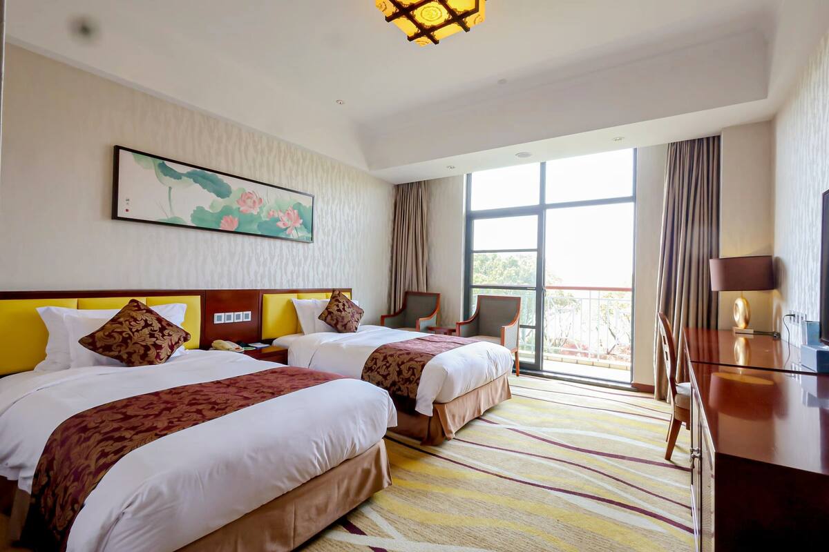 A hotel room with two beds, a desk, a chair, a large window, and a painting on the wall.