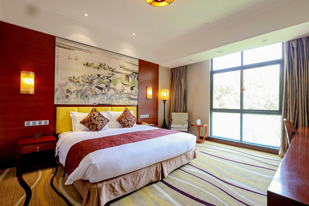 A hotel room with a large bed, a painting on the wall, a desk, and a window with curtains.