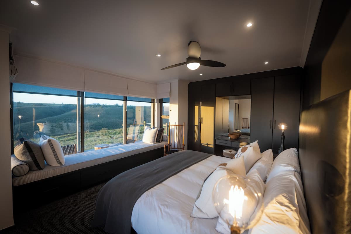 Un dormitorio con una cama grande, un rincón con asiento junto a la ventana, un ventilador de techo y vistas al exterior.