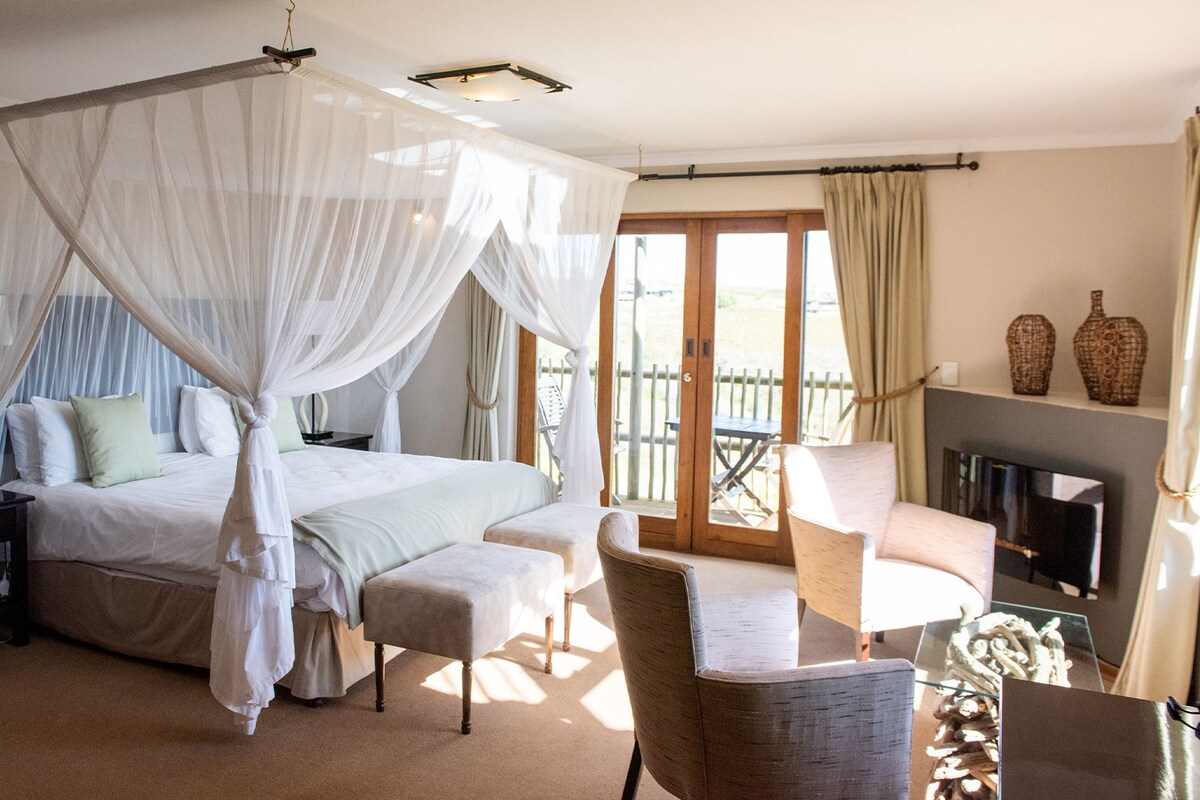Un dormitorio con cama con dosel, una zona de estar con una butaca y reposapiés, chimenea y balcón con vistas.