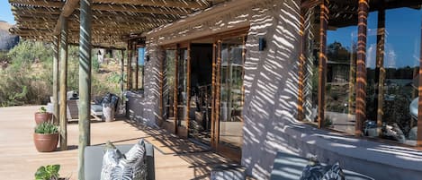 Un espacio de estar al aire libre con techo, bancos de madera y cojines, plantas en macetas y vistas del paisaje circundante.