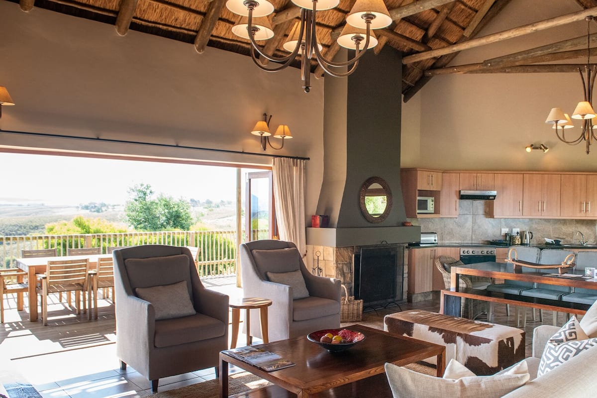 Amplia sala de estar con chimenea, mesa de comedor y asientos cómodos. Se observa una cocina con armarios de madera y electrodomésticos modernos al fondo.