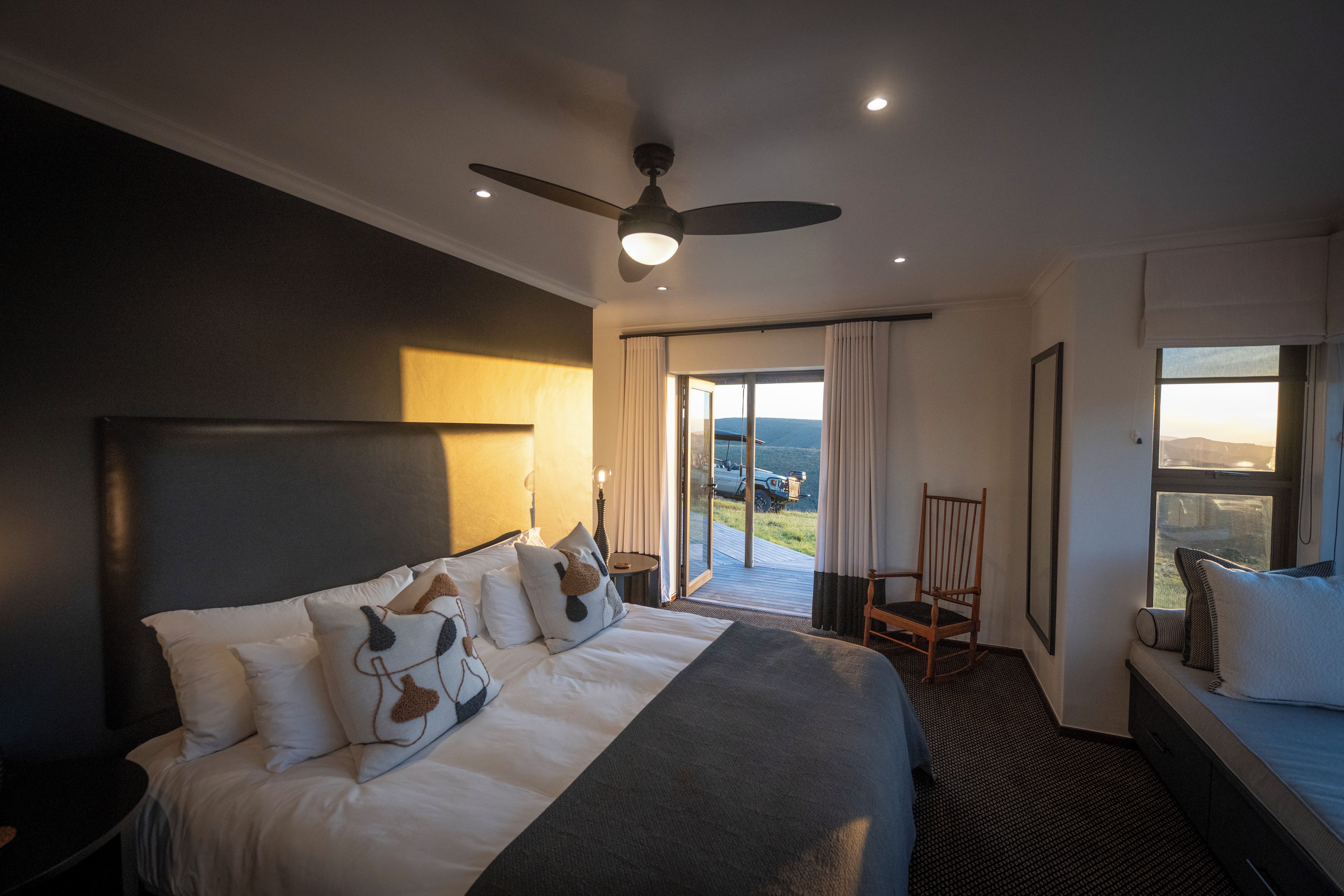 A hotel room with a large bed, a ceiling fan, a view of the outdoors, and a seating area by the window.