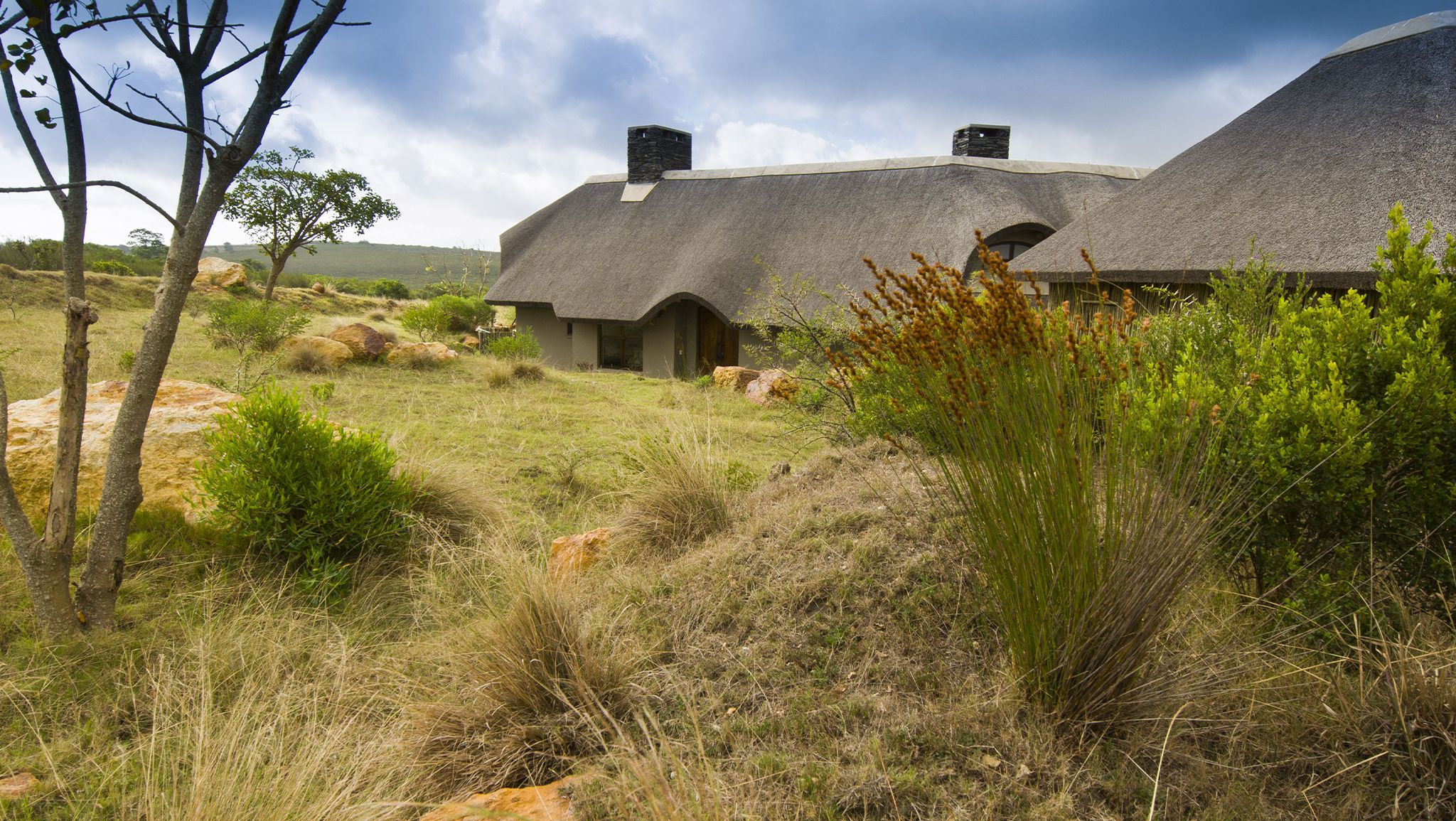 A thatched-roof house with a stone chimney, surrounded by natural vegetation and hills.