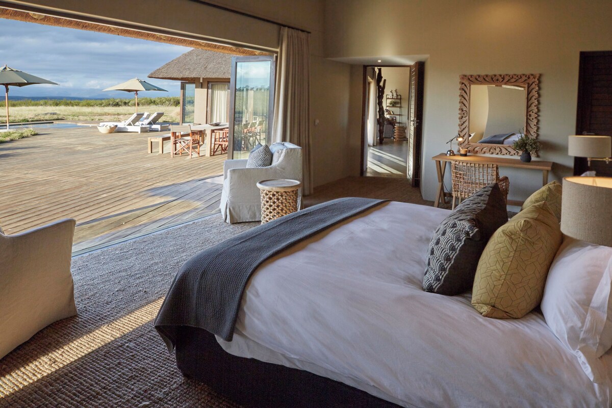 Habitación de hotel con una cama grande, vista a la playa, zona de comedor y sala de estar con sombrillas.