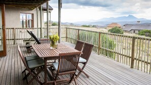 Una terraza de madera con una mesa, sillas, una planta en maceta y vistas a montañas distantes.
