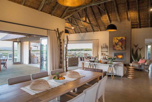A spacious living area with a wooden dining table, white sofas, and a fireplace. - Gondwana Game Reserve (Mossel Bay)