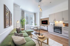 Ein modernes Wohnzimmer mit einem grünen Sofa, einem Holztisch und einer Küchenzeile mit weißen Schränken und einem Einbauofen.