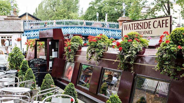Ett kafé vid vattnet med uteservering, en skylt som visar var det ligger i Little Venice, och en bro i bakgrunden.