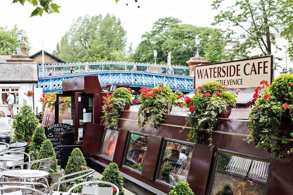 Ein Café am Wasser mit Außenplätzen, ein Schild, das den Standort in Little Venice angibt, und eine Brücke im Hintergrund.