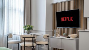 Ett modernt vardagsrum med en platt-TV som är monterad på väggen, ett runt matbord med stolar, en krukväxt och ett minimalistiskt avlastningsbord med dekorativa föremål.