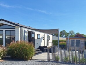 Ein Mobilheim mit einer Terrasse, einem eingezäunten Garten und einem kleinen Geräteschuppen.