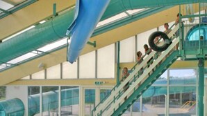 An indoor swimming pool with a water slide, a staircase, and a view of the outside. - CS 34 - Vakantiepark Callassande by Interhome (Callantsoog)