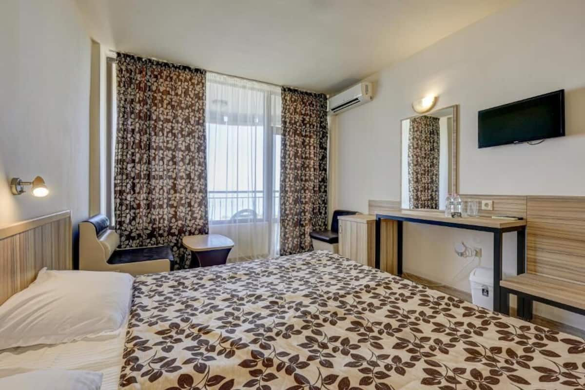 A hotel room with a bed, a desk, a chair, a TV, and an air conditioning unit.