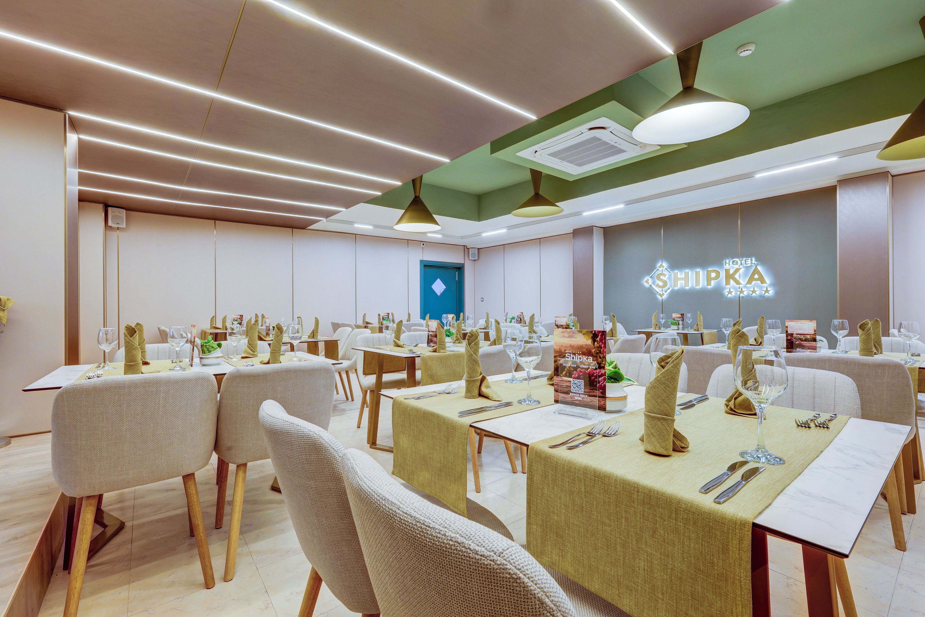 Une salle à manger d’hôtel avec plusieurs tables dressées pour les clients, une porte bien visible et un mur sur lequel est inscrit le nom de l’hôtel.