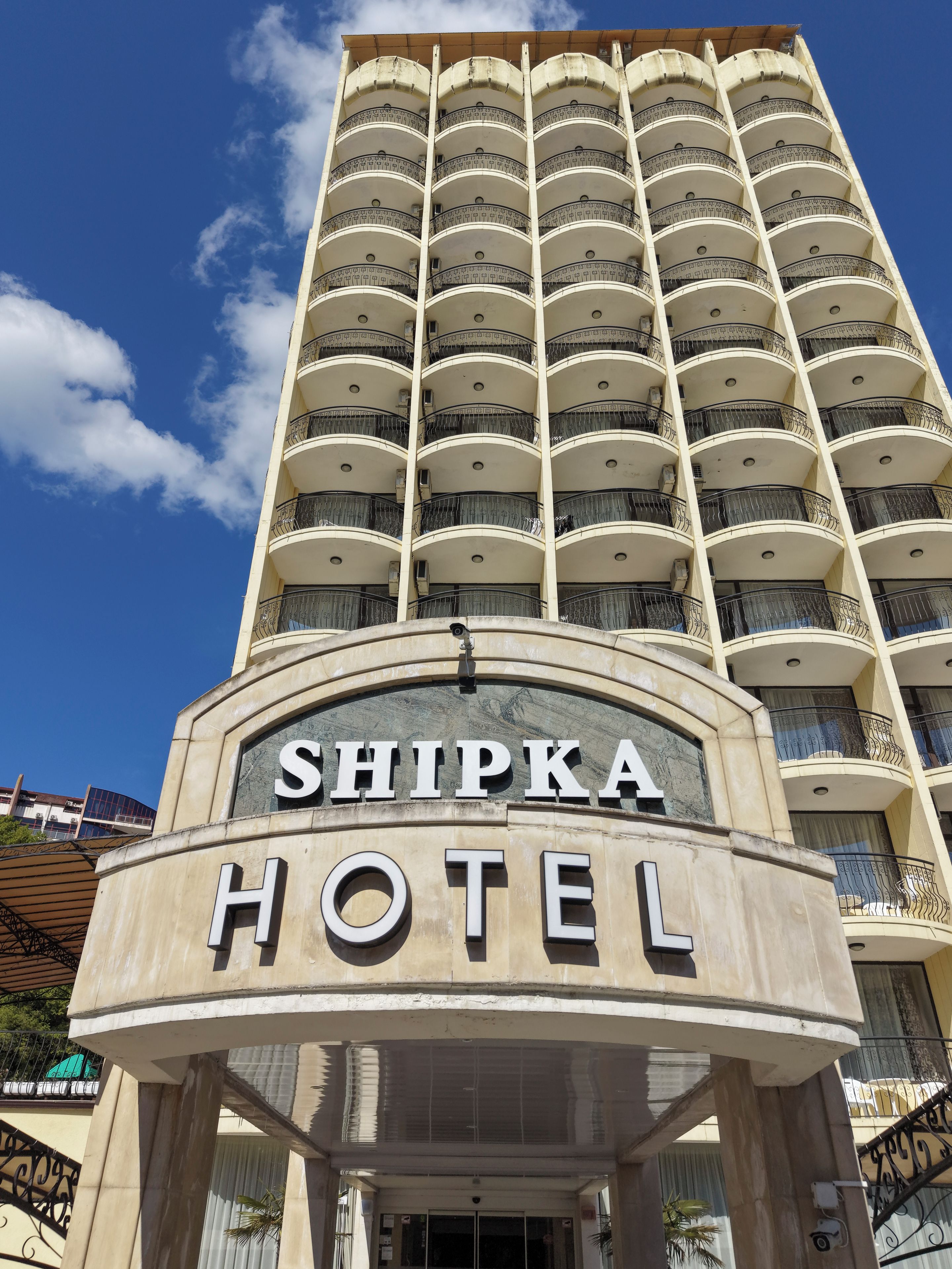 Un hotel con un letrero llamativo que dice 'SHIPKA HOTEL'. Tiene una estructura de varios pisos con balcones en cada planta.