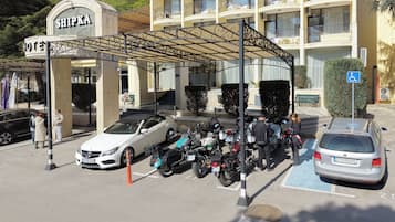 Entrada de hotel con zona de estacionamiento techada, un coche blanco y varias motocicletas aparcadas.
