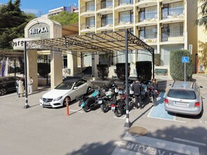Entrada de hotel con zona de estacionamiento techada, un coche blanco y varias motocicletas aparcadas.