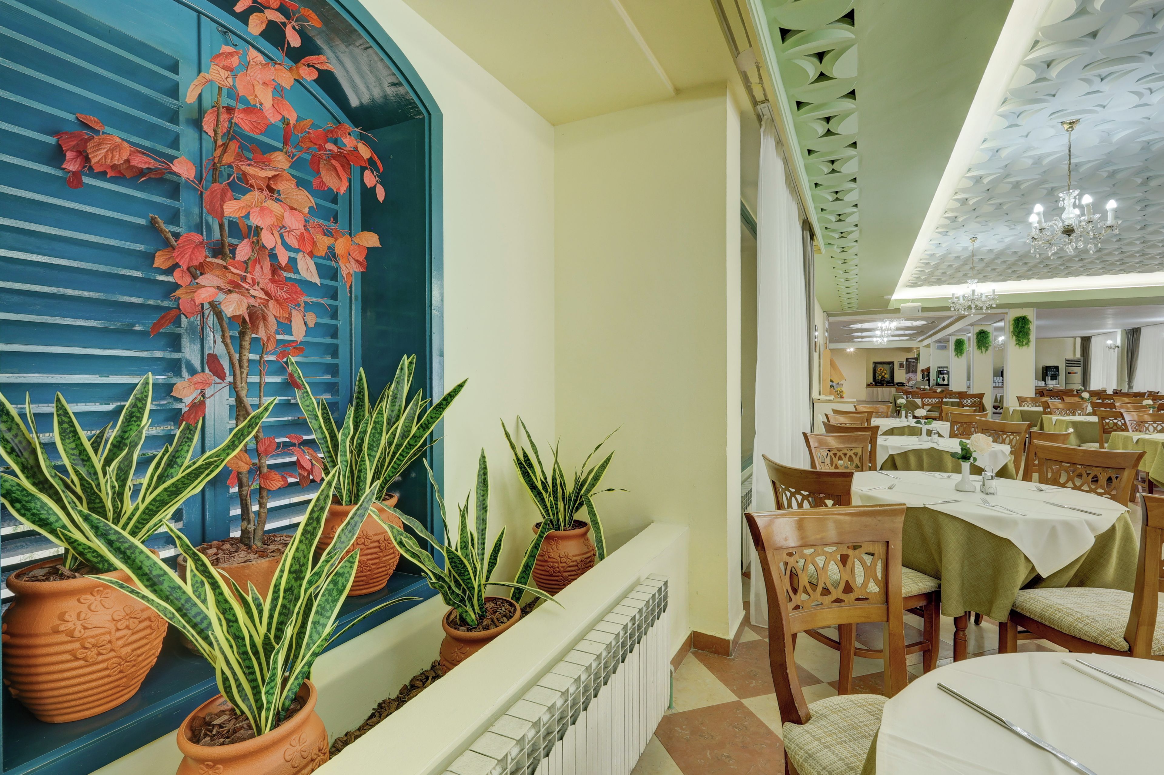 A dining area with tables set for service, potted plants, and a large window with red foliage.