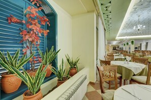 A dining area with tables set for service, potted plants, and a large window with red foliage. - Hotel Shipka All Inclusive (Varna)