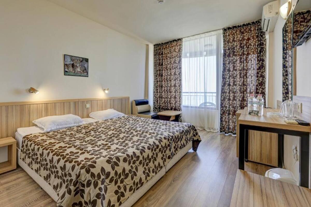 A hotel room with a bed, a desk, a chair, a small table, a picture on the wall, and a window with curtains.