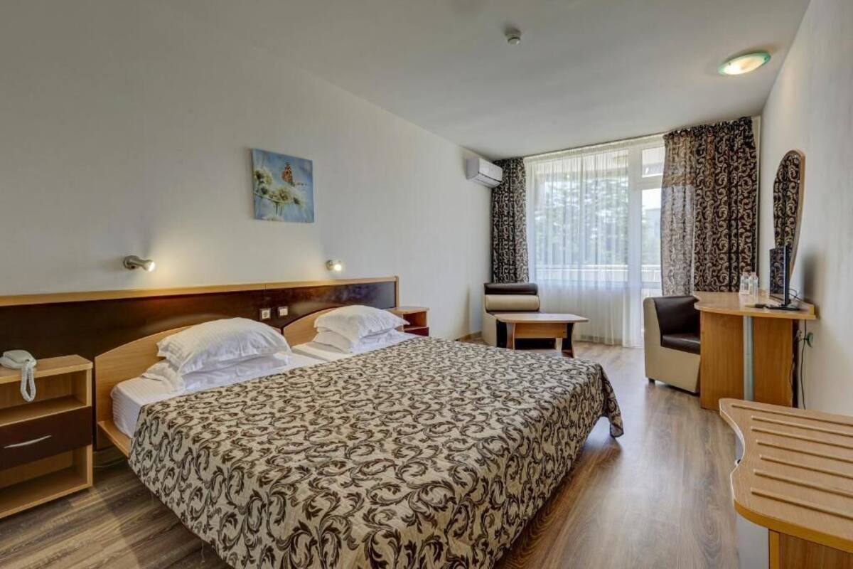 A hotel room with a bed, bedside table, chair, desk, and a painting on the wall.